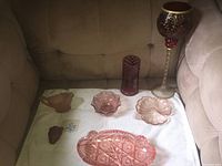 Seven pieces of vintage pink and red coloured glassware displayed on a beige fabric surface. Includes goblet, pitcher, bowls, vase, and trays.