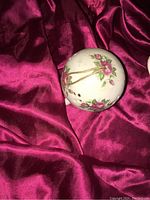 Porcelain round diffuser decorated with pink roses and a golden hanging cord on a pink silky fabric background.