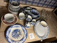 Full view of all dishes and serveware on counter including various stacked Blue Amari plates, bowls, cups, and serving platters plus contrasting pink floral bowl and clear glass cups.