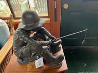 Side view showing boy sitting, fishing rod held in hands, wearing hat and boots, some chipping on statue