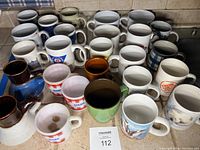 Photo showing 26 assorted coffee mugs in various colors, patterns, and logos on a kitchen counter.