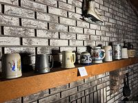 Side view of 9 beer steins arranged on a wooden mantel with a white painted brick background, showing the variety of mug shapes, colors and designs, including metallic and ceramic steins.