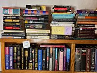 Image showing two wooden bookshelves filled with fiction books including authors like John Grisham, Herman Wouk, Sandra Brown, and various others. Books are both hardcover and paperback, arranged on shelves and stacked.