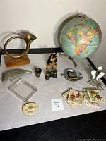 Full lot showing globe, brass horn, glass ashtray, binoculars, metal cup, carved stone bird, dog figurine, glass sculpture, and stamp booklets