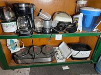 Photo showing assorted small kitchen appliances on a green shelf including coffee maker, toaster, blender parts, crockpot, and a pitcher.