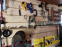 Workshop wall with various clamps (yellow and blue Quick-Grip clamps, metal C-clamps), drill press, radial arm saw blade, hand tools arranged on wood slat wall.