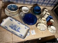 Top-down view of the assorted dinnerware including plates, bowls, cups, serving platter, and a blue glass cover. Various colors and patterns present.