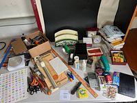 Wide view of various office and crafting supplies spread on a table including scissors, rulers, staplers, tapes, sticker sheets, and other small tools and materials