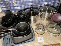 Overview of pots, pans, and glass cookware including stock pots, frying pans and colander, placed on kitchen counter against blue plaid wallpaper.
