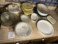 Full view of vintage Pyrex glass dishes including white casseroles with lids, clear glass bowls, and yellow-topped containers, arranged on countertop.