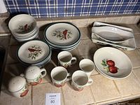 Top-down view of the red bird and apple dinnerware set showing plates, bowls, mugs, salt and pepper shakers, and serving dishes arranged on a tiled counter.