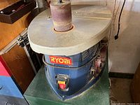 Photo showing a Ryobi belt sander with large sanding drum and attached sanding belt, blue body, green metal base, additional cylindrical sanding drums on side, and dust on surfaces.