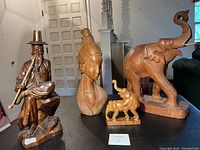 Four teak wood figures displayed on a table including a seated man playing a pipe, female bust, large elephant, and small elephant with riders.