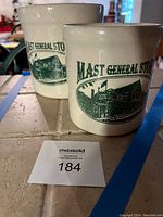 Photo showing two cream colored stoneware pots with green Mast General Store logos depicting a building and text.