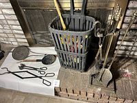 Photo of black metal basket, various black wrought iron fireplace tools including shovel, brush, poker, tongs, forks, and other implements laid out on fireplace hearth