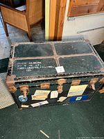Photo of the closed black vintage steamer trunk showing leather straps, metal hardware, corner protectors, stenciled address, and travel stickers.