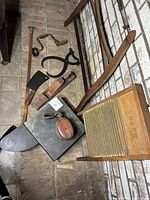 Overview photo showing vintage wooden washboard, hand drill, cleaver, wooden plane, tongs, and pulley alongside other assorted tools on brick surface.