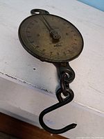 Angled side view of antique hanging weigh scale showing the brass dial, pointer, and iron hook beneath.