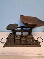 Overall view of cast iron balance scale with curved bowl on one side, flat platform on the other, and six weights displayed on the base