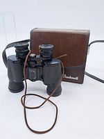 Photo showing Bushnell binoculars next to brown carrying case with strap and Bushnell logo on case.