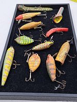 Photo showing the full set of 11 vintage wooden fishing lures arranged in a black display tray, displaying various shapes, colors, and hook configurations.