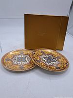 Two porcelain side plates displayed side by side with original gold-colored box behind them, showing full patterned surfaces