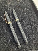 Two closed black Mabie Todd fountain pens with gold bands and clips resting on black fabric.