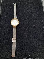 Full view of the wristwatch laid flat on a black surface showing silver mesh band and gold-tone round face.