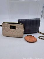 Three women's handbags and a small change purse on a white background. Beige Betsey Johnson handbag in front, gray clutch with chain strap behind on right, small round pleated orange purse in front right.