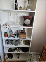 White shelving unit holding assorted vintage and antique decorative items, metal mugs, glass decanters, a Coca-Cola bottle, and various small items.