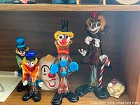 Front view of four Murano glass clown figurines along with two clown head figurines on a wooden shelf