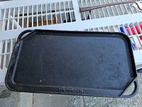 Top and angled views of the black rectangular KitchenAid cast iron griddle showing the cooking surface, integrated handles on both sides, a corner spout for grease pouring, and the KitchenAid embossing.