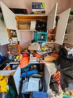 Wide shot showing white open cabinets filled with various items plus workbench area cluttered with boxes, storage bins, and tools.