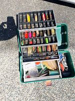 Open green metal fishing tackle box showing three tiered trays filled with assorted vintage fishing lures and hooks.