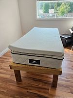 Side-angle photo showing twin mattress with foam top on custom wooden base near window and hardwood floor.