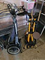 Two folding golf caddies, one black and one yellow, folded and standing upright in a storage area.