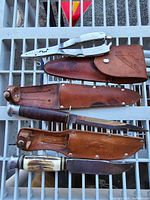 Photo showing two knives with leather sheaths and one metal cutter tool placed on metal grid surface.