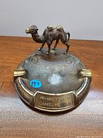 Front view showing camel figurine on rim, textured ashtray surface, cigarette rests, and name plaque