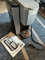 Photo of the Cuisinart coffee maker alongside the instruction manual, showing the front and side of the unit as well as the power cord and drip tray.