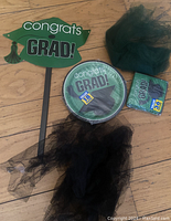 Photo showing a graduation cap photo prop, round paper plates with graduation theme, pack of 36 napkins, and two black tulle puffs for decoration on a wooden surface.