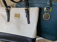 Two purses shown side by side, one cream and black with dark handles and gold square emblem, the other teal with gold lion head medallion hardware.