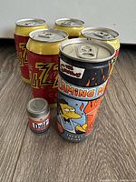 Lot showing four unopened Buzz Cola cans in the background and one unopened Flaming Moe Energy Drink can in front on a wood floor.