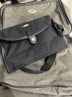 Photo showing the black Pierre Cardin flap bag placed on one of the olive green Eddie Bauer luggage pieces displaying their logos.