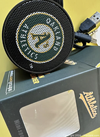 Close-up image of black mini speaker with perforated grille showing Oakland Athletics logo and USB and aux cables visible, packaged with branded box.