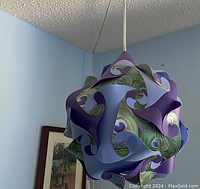 Photo showing the assembled decorative light fixture hanging from ceiling, made of interlocking curved panels in blue, purple and peacock feather pattern.