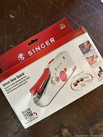 Box front and side of Singer Stitch Sew Quick handheld sewing machine showing product name, branding and images of included accessories and usage.