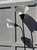 Standing floor lamp with two frosted white glass shades, one large upward-facing and one smaller flexible reading lamp, photographed outdoors against a grey wooden background in daylight.