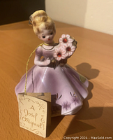 Photo showing porcelain figurine of a lady in a purple gown holding purple flowers, with original Josef Originals label attached by a string.