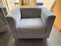 Front view of a gray upholstered boxy armchair with cushioned seat and armrests.