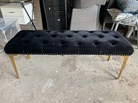 Front angled view of the bench showing tufted black fabric and gold metal legs on a concrete floor.